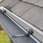 Close-up of a black mesh gutter guard installed on a home with gray asphalt shingle roof, showing the screen and mounting brackets.