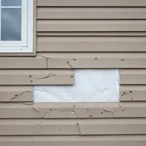 Home | Teague Roofing Plus 57 Hail-cracked tan vinyl siding with two missing panels exposing white house wrap stapled to the wall behind it